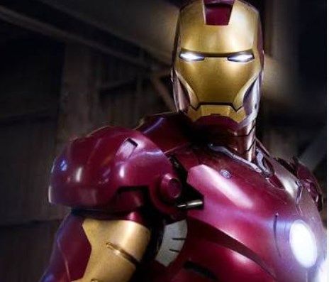 Iron Man w czerwono-złotej zbroi, stoi w ciemnym pomieszczeniu, emitując niebieskie światło z oczu.